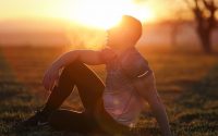 Muscular man sits in rural field at sunset, exhaling visible breath.